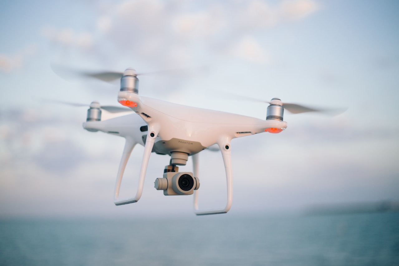 White quadcopter drone with camera gimbal hovering against a pale blue sky with soft clouds and hazy water below