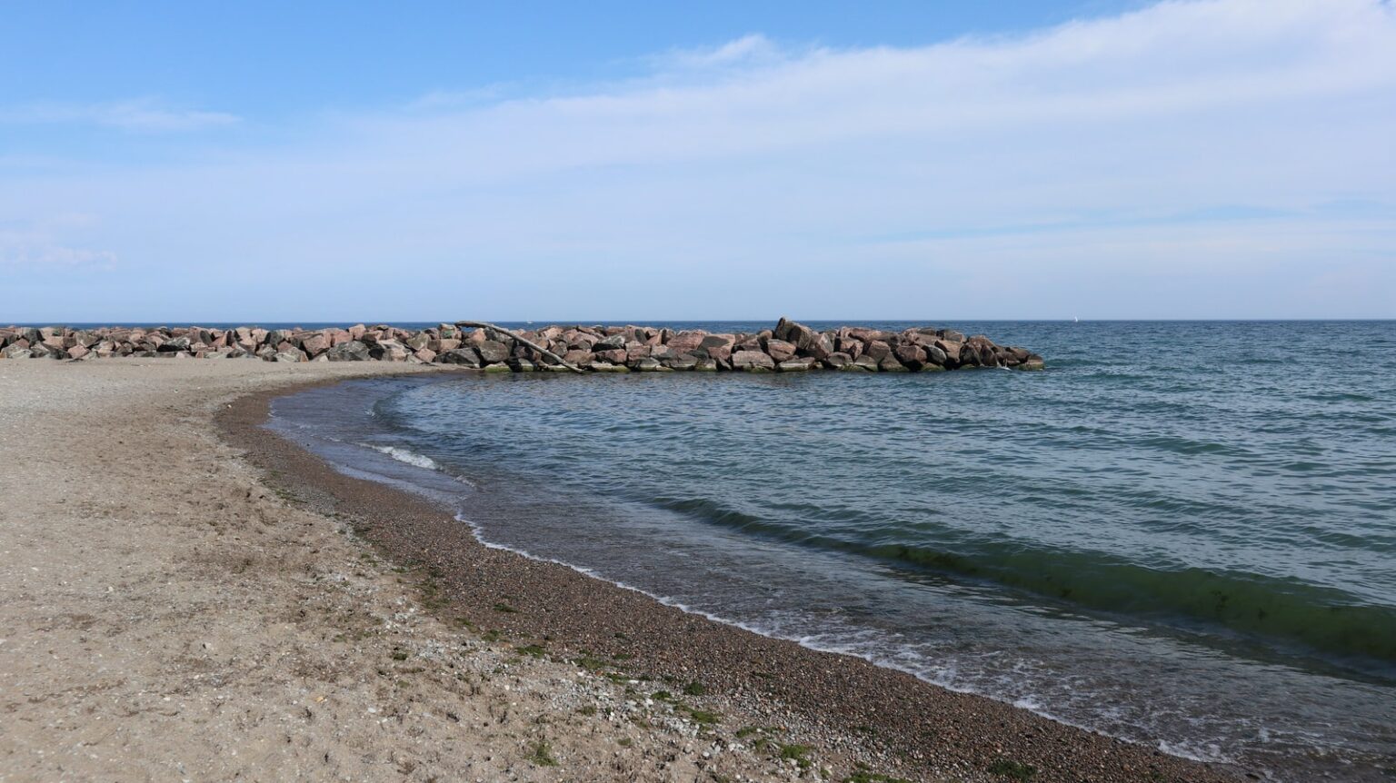 Aerial overhead flat-lay photograph of a curving pebble and sand shoreline meeting a calm pale blue sea, with a low rock breakwater extending into the water under a soft hazy sky; muted blues, sands, and greys, calm Mediterranean mood.