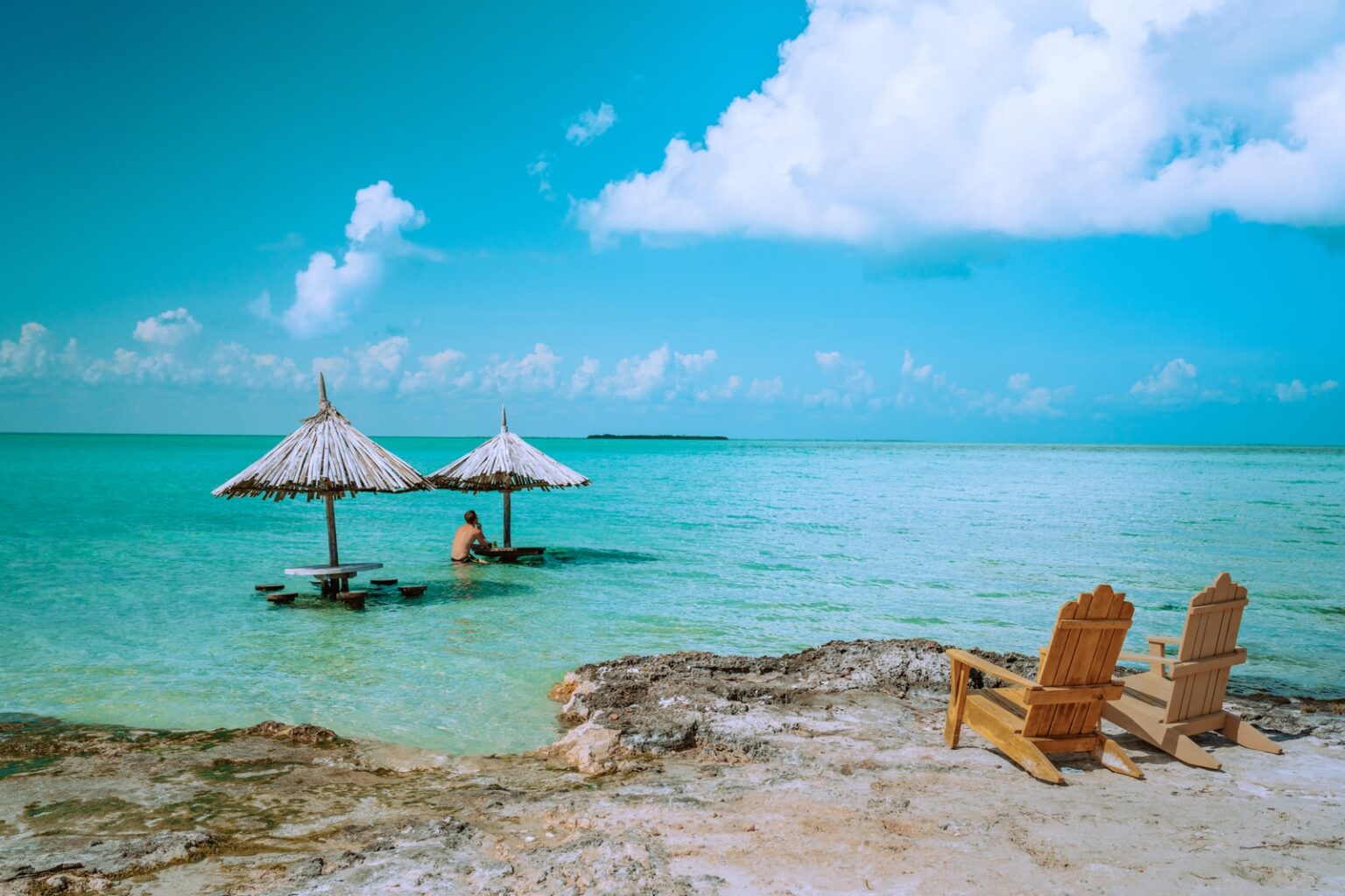 Two wooden beach chairs on a rocky shoreline overlooking a turquoise sea with thatched umbrellas and a wading figure; bright sky with soft clouds, in warm coastal blue and sand tones.